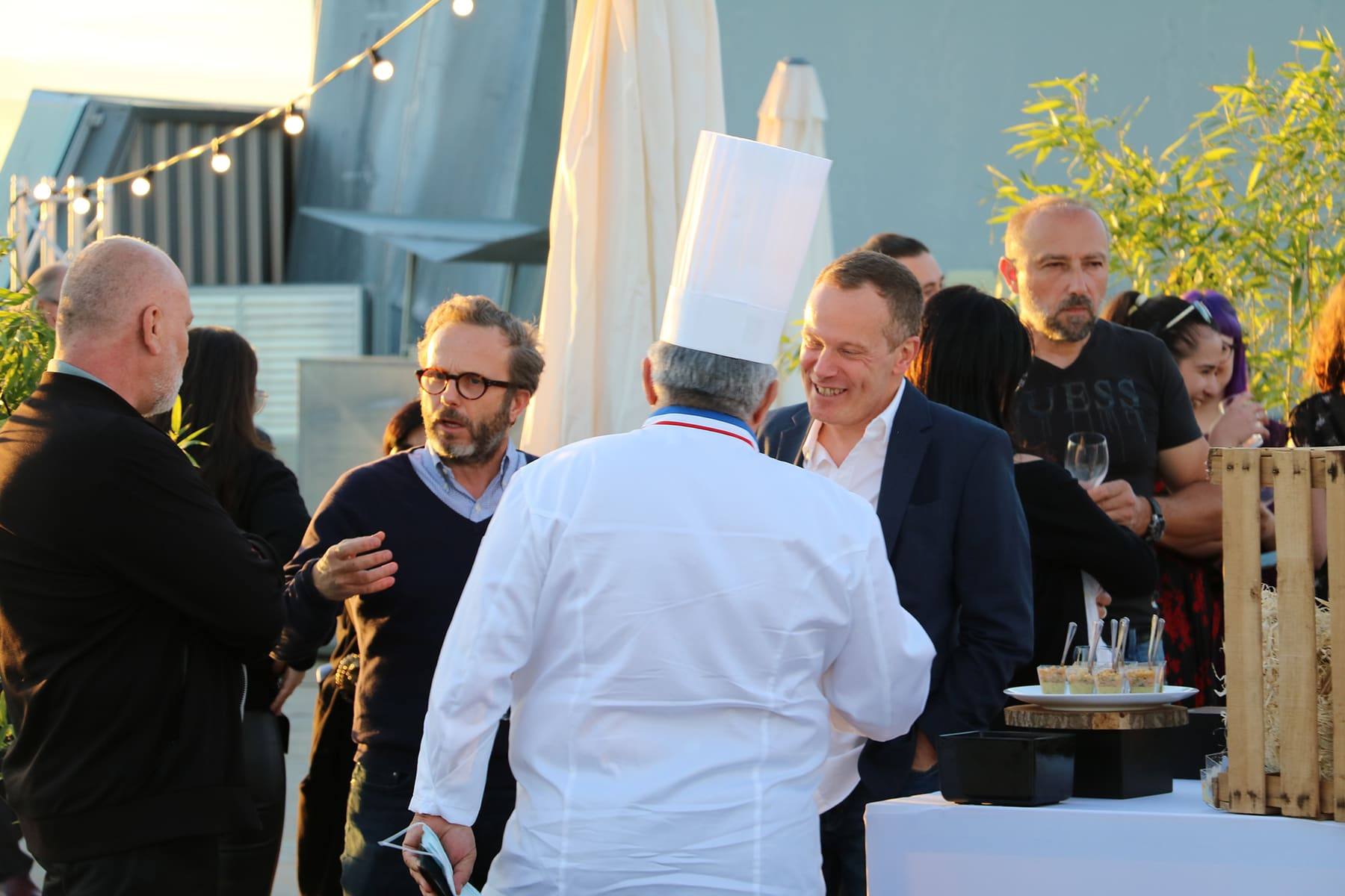 Toque-wearing chef greeting guests in suits at a high cocktail table on the rooftop