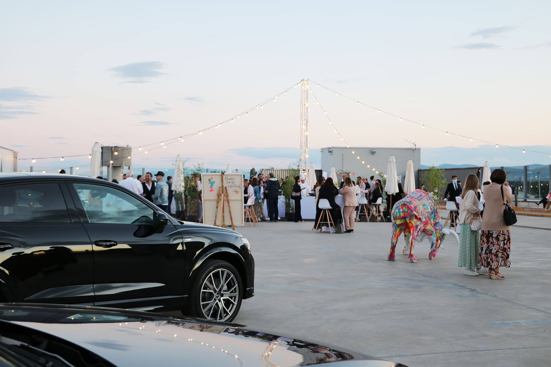 Painted bull sculpture and parked Audis framing the rooftop cocktail crowd at twilight