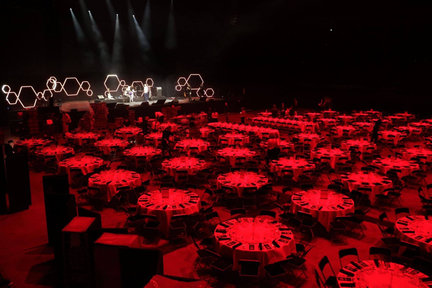Banquet floor of round tables under deep red light as the band performs on the hexagon-lit stage