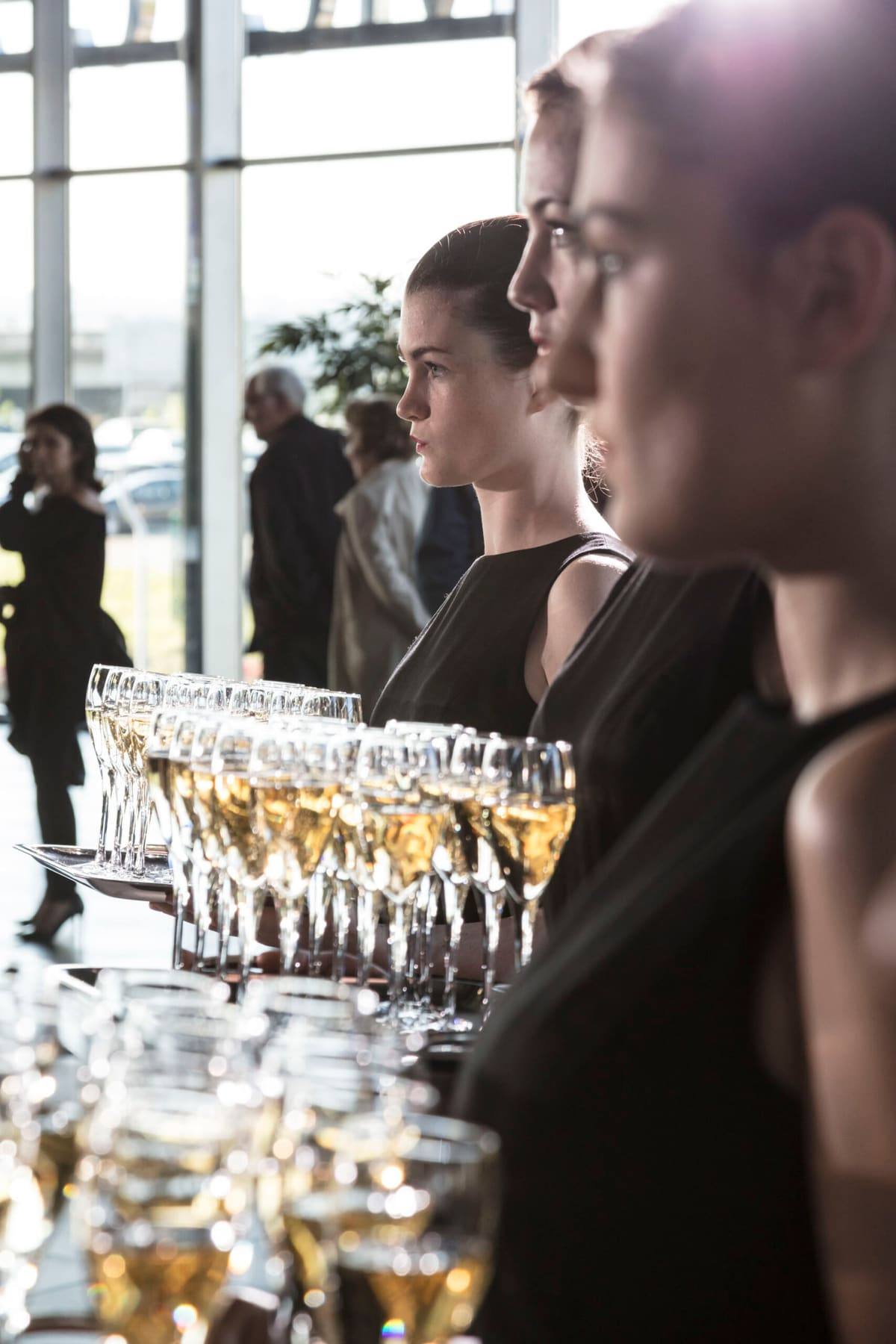 Hostesses in black holding trays of champagne flutes in the sunlit glass atrium