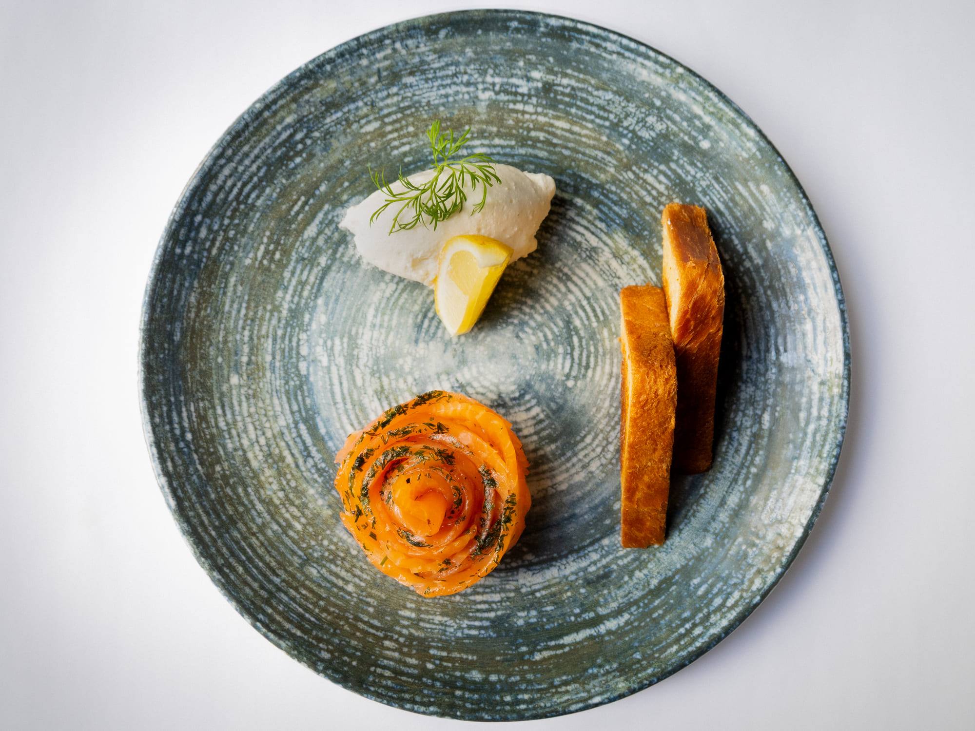 Assiette de grès vue de dessus, fines tranches de poisson mariné en rosace, quenelle de crème et pain grillé