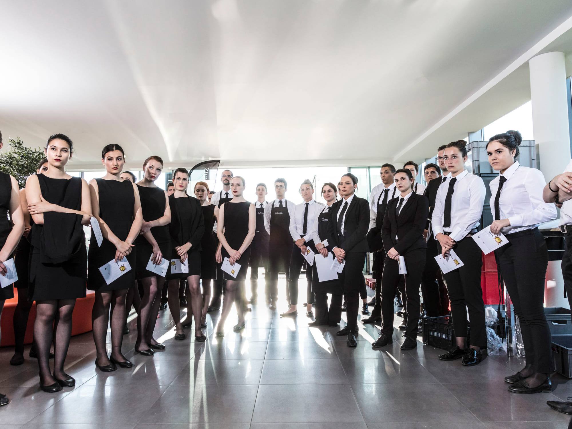 A long line of service staff holding briefing sheets in a glass-walled hall