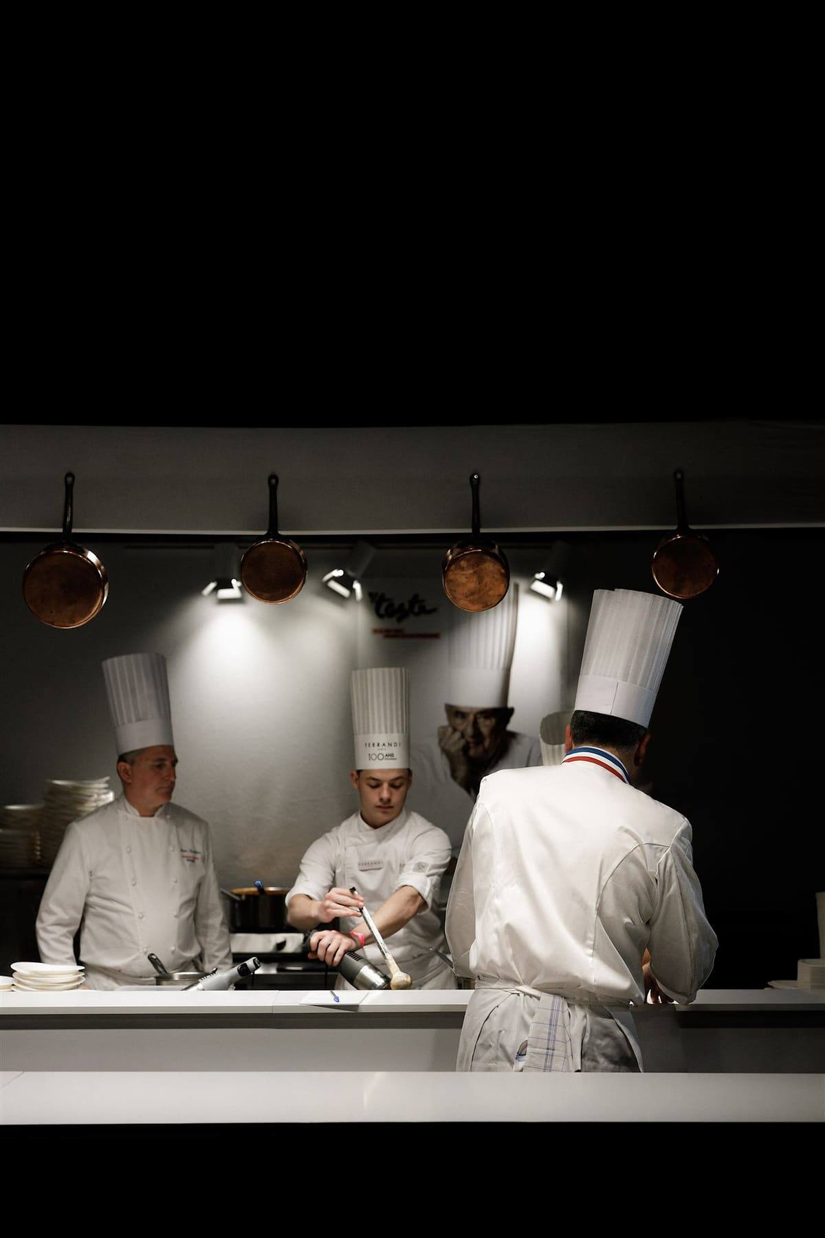 The Bocuse open-kitchen stand at Taste of Paris, chefs at work beneath hanging copper pans